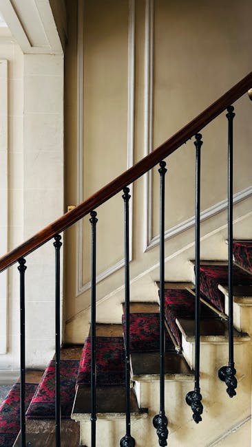 An indoor staircase with wooden handrails and black metal balusters ascending from a ground floor to an upper level in a residential property. The staircase is partially covered with a red patterned c