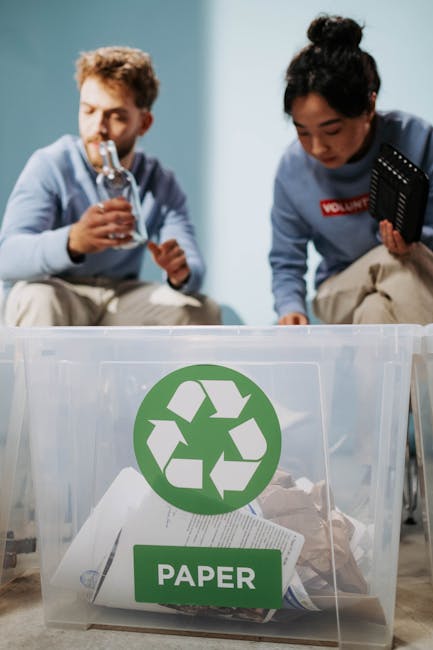 Two volunteers are situated inside a home during a house removal process, with a clear plastic recycling bin in the foreground containing crumpled paper and other packaging waste, marked with a green 