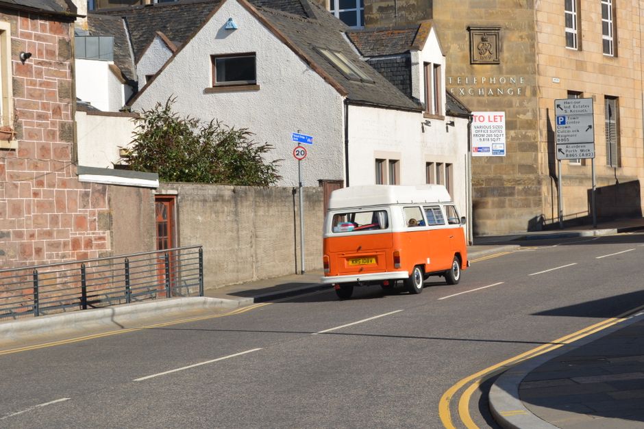 A white commercial van parked on a grassy area beside a residential building with red brick walls and window boxes. The van's rear wheel and part of its side panel are visible, with the side panel sho