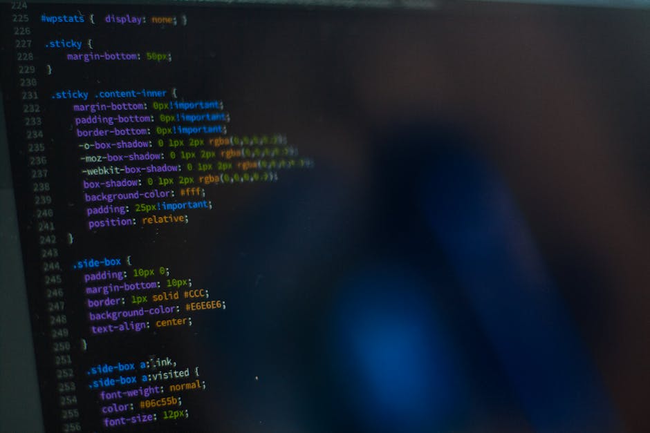 Close-up image of a person’s hands operating a laptop on a dark table, with the screen displaying lines of coding and software development interface. The background shows a blurred dark setting, emp